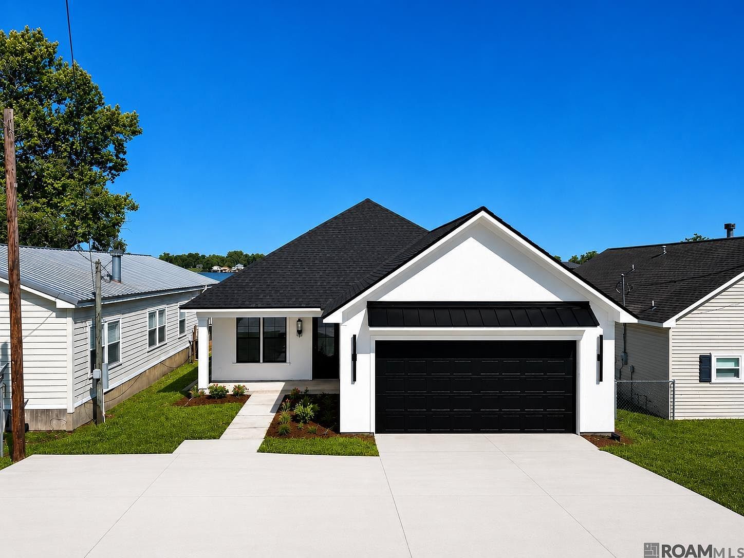 Front elevation of 8749 Island Rd showing a modern white cottage with a black metal-accent gable, two-car garage, and clean concrete driveway under a Louisiana blue sky.