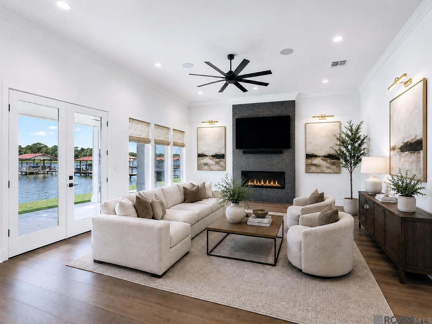 Great room with a charcoal-tile gas fireplace beneath a wall-mounted television, large windows opening to the boat dock and False River beyond, and a low-slung cream sectional centered on the hearth.