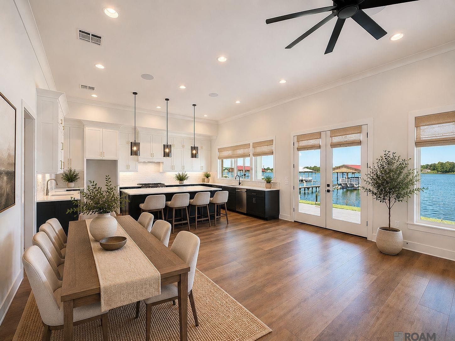 Open kitchen with a large island, pendant lighting, custom cabinetry to the ceiling, a 36-inch gas range, and the dining table set against full glass doors looking out over False River.