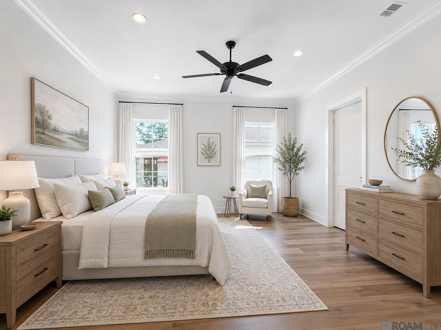 Primary bedroom suite with a tray ceiling, a wide window flooded with morning light, a cream upholstered headboard, and warm oak floors running edge to edge.